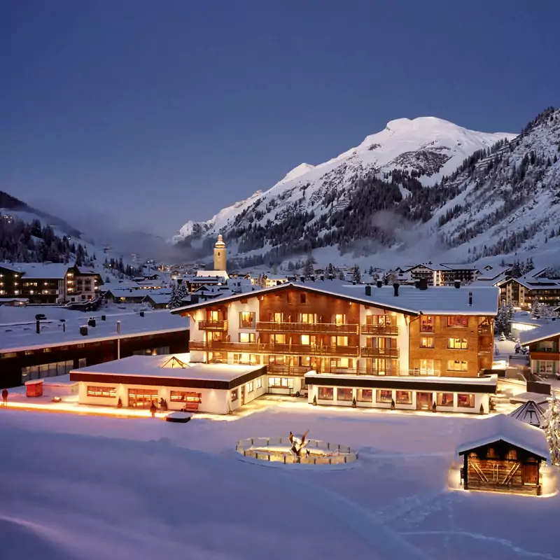 Hotel Auriga in Lech am Arlberg during a snowy winter evening with illuminated windows and surrounding snow-covered mountains.