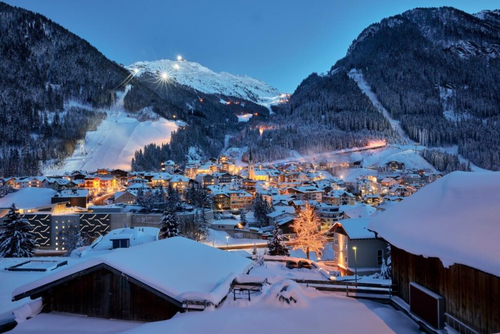 A picturesque night view of Ischgl town, illuminated with warm lights, surrounded by snow-covered mountains and ski slopes.